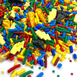May include: A colorful assortment of sprinkles in various shapes and sizes. The mix includes yellow lightning bolts, gold stars, black boots, and orange horns, along with red, green, blue, and yellow sprinkles. The sprinkles are scattered on a white surface.