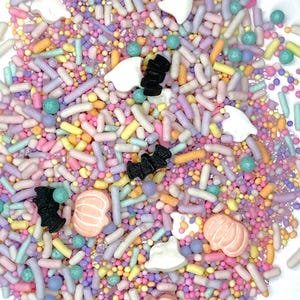 May include: A colorful mix of Halloween-themed sprinkles. The mix includes pastel-colored sprinkles, small round sprinkles, and decorative shapes like ghosts, bats, and pumpkins. The sprinkles are in shades of pink, yellow, blue, and purple.