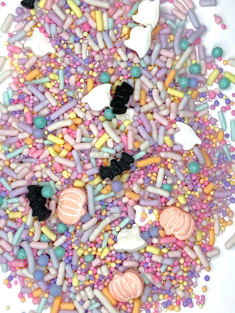 May include: A colorful mix of Halloween-themed sprinkles. The mix includes pastel-colored sprinkles, small round sprinkles, and decorative shapes like ghosts, bats, and pumpkins. The sprinkles are in shades of pink, yellow, blue, and purple.