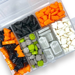 Könnte beinhalten: Ein durchsichtiger Plastikbehälter mit sechs Fächern, gefüllt mit Halloween-Streuseln. Die Streusel sind in verschiedenen Farben, darunter Orange, Schwarz, Weiß, Grün und Grau. Einige der Streusel sind in Form von Geistern, Fledermäusen, Kürbissen und Grabsteinen. Der Behälter ist geöffnet und die Streusel sind sichtbar.