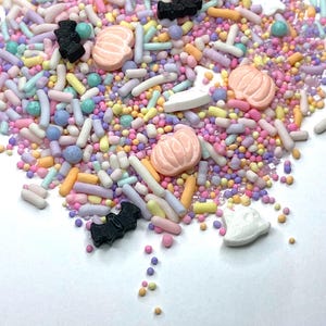 May include: A colorful mix of sprinkles with pastel colors, including pink, yellow, blue, and purple. The sprinkles include small round balls, long rods, and shapes like pumpkins, bats, and ghosts. These are likely for decorating baked goods.