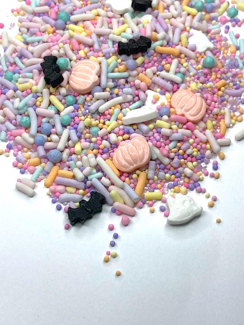 May include: A colorful mix of sprinkles with pastel colors, including pink, yellow, blue, and purple. The sprinkles include small round balls, long rods, and shapes like pumpkins, bats, and ghosts. These are likely for decorating baked goods.