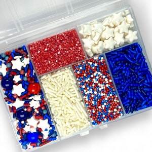 Op de afbeelding: Een doorzichtige plastic container met zes compartimenten gevuld met rode, witte en blauwe sprinkles. De sprinkles omvatten sterren, kleine balletjes en lange, dunne sprinkles.