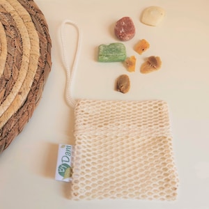 May include: A white mesh bag with a drawstring closure and a white tag that says "By Dani". The bag is on a white surface with a variety of colourful stones and soap bars.