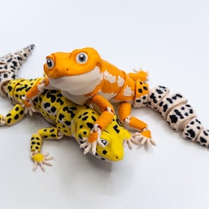 Puede incluir: Dos geckos de juguete, uno naranja y blanco con manchas negras, el otro amarillo con manchas negras, están posados juntos sobre un fondo blanco. El gecko naranja está encima del gecko amarillo.