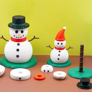 Op de afbeelding: Een verzameling sneeuwman-thema decoraties. Twee witte sneeuwmannen met zwarte knoopogen en kolenmonden zijn afgebeeld, een met een hoge hoed en de andere met een rode hoed. Extra stukken zijn hoeden, sjaals en donutvormige bases.