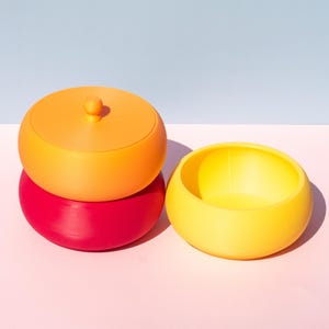 May include: Three nesting bowls in shades of orange, yellow, and red. The top bowl has a lid.