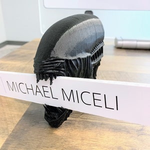 May include: A black 3D printed model of an alien head with a white sign that reads "MICHAEL MICELI" in front of it.