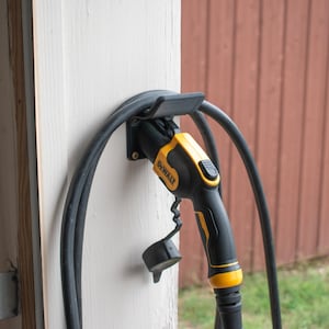 May include: A black and yellow electric vehicle charger mounted on a white wall. The charger has a black cable and a black holster. The brand name "DEWALT" is visible on the charger. The background includes a garage door and a wooden fence.