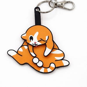 May include: Orange and white cat-shaped keychain with a black strap, silver keyring, and clasp. The cat is lying down with its eyes closed and tongue sticking out. The keychain is a fun accessory.