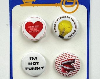 May include: A set of four round button badges with different designs. The first badge has a red heart with the text "SUCKER FOR LOOKS". The second badge has a yellow lightbulb with the text "WOULD YOU LEAVE ON THE LIGHT FOR ME?" The third badge has white text on a white background that says "I'M NOT FUNNY". The fourth badge has a red and white image of a chattering teeth toy with the text "29292929" repeated around the edge.