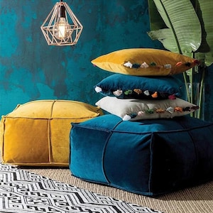 Puede incluir: Dos pufs, uno amarillo y otro azul, con una pila de cuatro almohadas decorativas con borlas. Las almohadas son en tonos de amarillo, blanco y azul. Los pufs y las almohadas están sobre una alfombra con estampado blanco y negro.