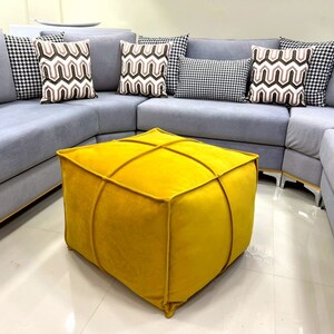 Puede incluir: Un puf cuadrado amarillo con un diseño capitoné se encuentra frente a un sofá seccional gris con almohadas decorativas estampadas.