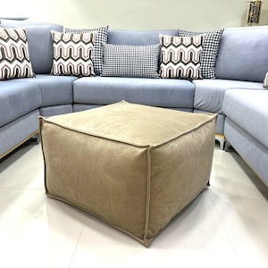 Puede incluir: Un puf cuadrado beige con una cubierta de tela suave y mullida. El puf se coloca frente a un sofá seccional azul claro con almohadas decorativas estampadas.
