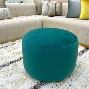 Puede incluir: Un puf redondo de color verde azulado con una textura suave y aterciopelada. El puf está sobre una alfombra con estampado blanco y marrón.