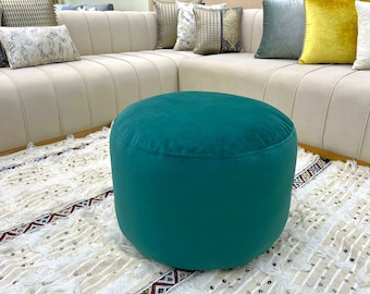 Puede incluir: Un puf redondo de color verde azulado con una textura suave y aterciopelada. El puf está sobre una alfombra con estampado blanco y marrón.