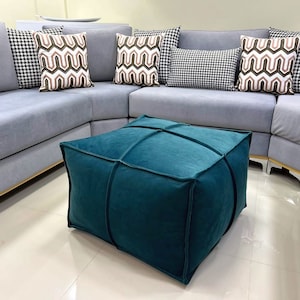 Puede incluir: Un puf cuadrado azul turquesa con un diseño capitoné se encuentra delante de un sofá seccional gris con almohadas decorativas estampadas.