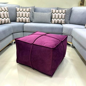 Puede incluir: Un puf cuadrado de terciopelo morado con un diseño capitoné. El puf está situado delante de un sofá seccional gris claro con almohadas decorativas estampadas.