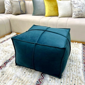 Puede incluir: Un otomán cuadrado turquesa con detalles de costura se encuentra sobre una alfombra estampada. El otomán está rodeado por un sofá color crema con varias almohadas decorativas en tonos grises, amarillos y blancos.