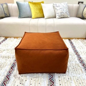 Puede incluir: Un puf cuadrado marrón con una alfombra de flecos blancos debajo. El puf está hecho de una tela suave y mullida.