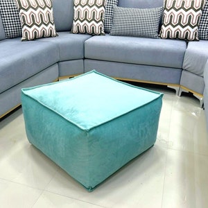 Puede incluir: Un otomán cuadrado de terciopelo turquesa se encuentra sobre un suelo embaldosado frente a un sofá seccional azul claro. Cojines decorativos con estampados geométricos y pata de gallo adornan el sofá. El otomán tiene forma de cubo.