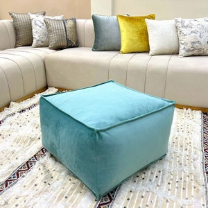 Puede incluir: Un puf cuadrado de color azul claro sobre un fondo blanco con una alfombra de patrón blanco y rojo. El puf está hecho de una tela suave y mullida.