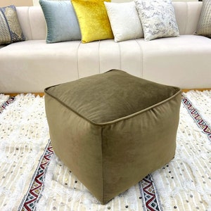 Puede incluir: Un puf cuadrado de terciopelo marrón se encuentra sobre una alfombra blanca y beige con flecos. El puf está delante de un sofá blanco con cojines.