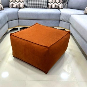 Puede incluir: Un otomano cuadrado marrón con un sofá seccional gris claro en el fondo.