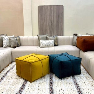 Puede incluir: Un sofá seccional blanco con una variedad de almohadas, incluyendo una almohada estampada con detalles dorados. Tres otomanas cuadradas en diferentes colores, amarillo, azul y marrón, se colocan frente al sofá. El sofá y las otomanas están sobre una alfombra blanca con un patrón geométrico.