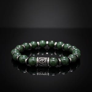 May include: A green beaded bracelet with silver-colored accents. The bracelet features round, dark green beads and silver-colored spacers. A central cylindrical bead with a Celtic knot design adds detail to the bracelet.