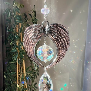 May include: A silver angel wing crystal suncatcher with a teardrop crystal and a round crystal hanging from it. The suncatcher is hanging from a chain.