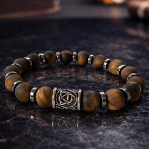 May include: A beaded bracelet featuring round, matte brown tiger eye beads alternating with small, polished silver-toned metal spacers. A central cylindrical silver-toned metal bead displays an embossed triquetra knot design.
