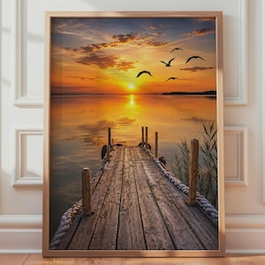 May include: A framed wall art print featuring a wooden pier extending into a calm lake at sunset. The sky is filled with vibrant orange and yellow hues, with silhouettes of birds flying above. The water reflects the golden sun, and reeds grow nearby.