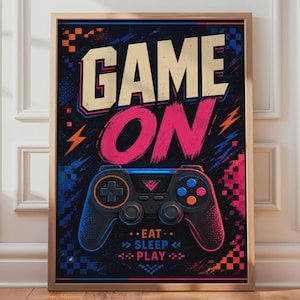 May include: A framed poster featuring the text GAME ON in large beige and pink letters above a black video game controller. Below the controller, the text EAT SLEEP PLAY is displayed in blue and pink. The background has blue and orange accents.