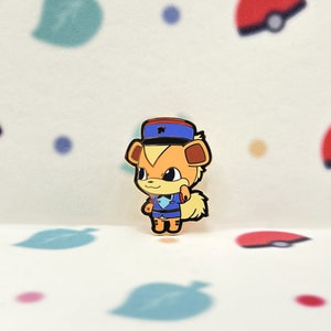 Puede incluir: Un pin de dibujos animados con un personaje canino naranja y marrón con uniforme y sombrero azules con una raya roja. El pin tiene una pequeña cola esponjosa. El fondo es blanco con formas coloridas.