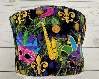 May include: A black scrub hat with a Mardi Gras theme. The hat features a purple, green, and gold design with a saxophone, masks, and fleur-de-lis.