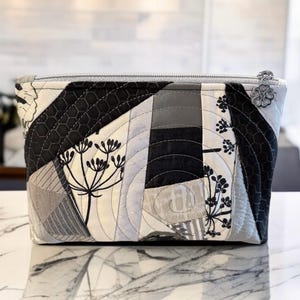 May include: A quilted cosmetic bag in a patchwork design of grey, black, and white fabrics. It has a silver zip and a floral zip pull. The bag features floral and honeycomb patterns, with a geometric design.