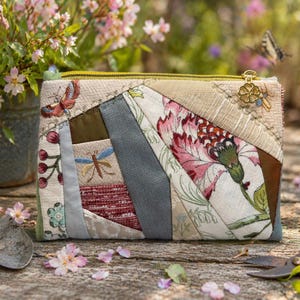 May include: A patchwork zippered pouch with floral and insect embroidery. The design features a mix of fabrics in various colors, including pink, green, and brown. A gold-colored zipper and charm add detail. The pouch is set on a wooden surface with gardening tools and flowers.