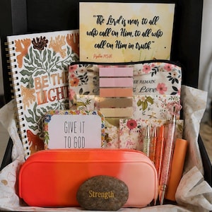 May include: A gift box containing a spiral notebook with the text BE THE LIGHT, a card with the text The Lord is near to all who call on Him, to all who call on Him in truth, a floral pouch, a card with GIVE IT TO GOD, pens, a stone with Strength, and an orange case.