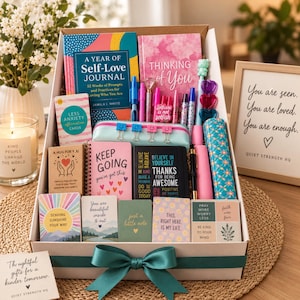 May include: A gift box containing a journal titled A Year of Self-Love Journal, a Thinking of You notebook, various pens, a pencil case with binder clips, affirmation cards, and small notebooks with text including Keep Going and Believe in Yourself.