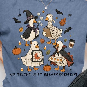 Może przedstawiać: Niebieski t-shirt z grafiką przedstawiającą cztery kaczki w kostiumach na Halloween, w tym czarownicę, mumię, ducha i wampira. Wzór zawiera dynie, nietoperze, liście oraz czarny napis NO TRICKS JUST REINFORCEMENT.