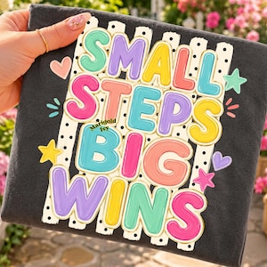 以下が含まれることがあります： 「SMALL STEPS BIG WINS」というバブルレターのテキストが描かれたカラフルなグラフィック付きのダークグレーのTシャツ。デザインには、黒い縦縞とドットが入った白い背景の上に、パステルカラーのハートと星が配置されています。