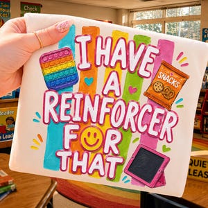 May include: A white rectangular pouch featuring the colorful text I HAVE A REINFORCER FOR THAT. The design includes a rainbow pop-it toy, a bag of snacks, a yellow smiley face, and a pink tablet, all set against vertical rainbow stripes.