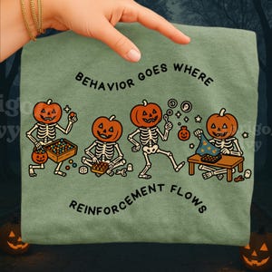 May include: Sage green t-shirt with a Halloween-themed graphic. The design features four skeleton figures with pumpkin heads, engaged in various activities. The text "BEHAVIOR GOES WHERE REINFORCEMENT FLOWS" is printed in a circular pattern.