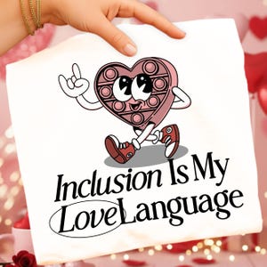 May include: White t-shirt featuring a cartoon heart-shaped pop-it toy with arms, legs, and red sneakers. The text "Inclusion Is My Love Language" is printed below the graphic in a stylish font.