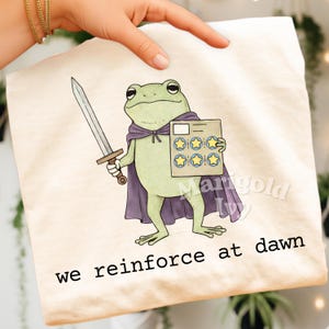 May include: A light beige t-shirt featuring a cartoon frog warrior with a sword and cape. The frog holds a card with star ratings. The text "we reinforce at dawn" is printed at the bottom.