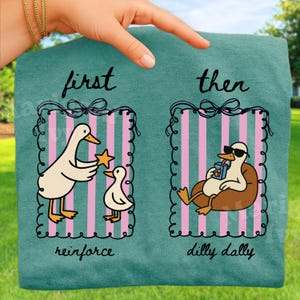 May include: Teal-colored t-shirt with a whimsical duck design. The shirt features two panels: one with a duck giving a star to a smaller duck, and the other with a duck relaxing. Text reads "first, then, reinforce, dilly dally."