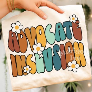 May include: Cream-colored t-shirt with the words "ADVOCATE INCLUSION" in a retro font, accented with daisy graphics. The text is in shades of brown, orange, teal, and yellow. The shirt is held by a person.