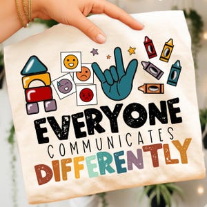 May include: Cream-colored t-shirt with the message "EVERYONE COMMUNICATES DIFFERENTLY" in black and colorful letters. The shirt features illustrations of building blocks, emojis, a hand gesture, and crayons.