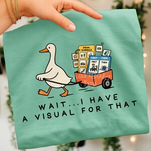 May include: A light teal t-shirt featuring a cartoon duck pulling a red wagon filled with visual schedule icons. The shirt has the text "WAIT... I HAVE A VISUAL FOR THAT" in black.